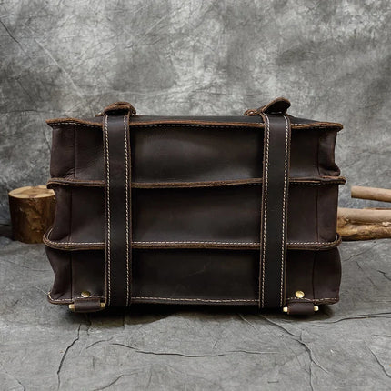 The Leather Business Travel Briefcase Backpack, crafted from dark brown genuine cowhide leather and featuring multiple compartments with beige stitching, is displayed against a textured gray background. This robust and stylish bag includes two handles and brass rivets at the bottom corners.