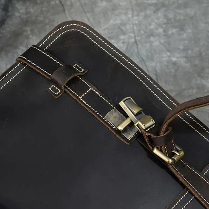 Close-up of the Leather Business Travel Briefcase Backpack in dark brown cowhide leather, highlighting a section of the flap adorned with white stitching, a brass buckle, and a leather strap loop. Made from genuine cowhide leather, it comes with a lifetime warranty. The background features a textured gray surface.