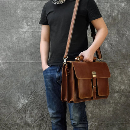 A person dressed in a black T-shirt and blue jeans holds the Leather 16" Business Briefcase, made of genuine cowhide leather with an adjustable strap, while standing against a textured grey backdrop. The bag features multiple compartments and brass hardware. The person's face is not visible in the image.