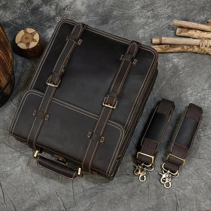 A dark brown genuine cowhide leather bag with light stitching, featuring two front straps and brass buckles, rests on a grey textured surface. Next to the Leather Business Travel Briefcase Backpack lie two detachable shoulder straps, both adorned with matching brass hardware. Sticks and a wooden log are visible in the background.
