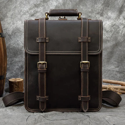 A dark brown Leather Business Travel Briefcase Backpack, made from genuine cowhide leather with white stitching and brass hardware, featuring a top handle and two front buckle straps, stands upright against a rustic background with wooden logs and tools.