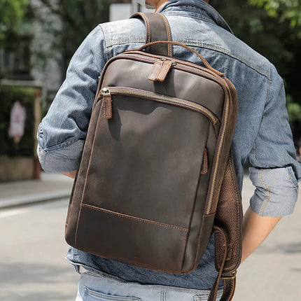 A person sporting a denim jacket strolls down the street with the Vintage Leather Daypack slung over one shoulder, set against a backdrop of trees and a building.