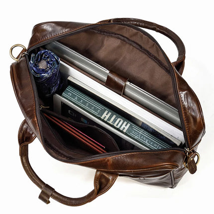 The Leather 16" Briefcase, crafted from genuine cow leather in a rich brown hue, opens to reveal an organized interior containing a folded umbrella, a securely strapped closed laptop, a book, a notebook, a red wallet, and several documents. This professional briefcase is designed with dual handles and an adjustable shoulder strap for added convenience.