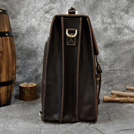 A vintage-style, dark brown Leather Business Travel Briefcase Backpack, crafted from genuine cowhide leather, is seen from its side, showcasing its sturdy construction and stitching. The background features a textured gray wall with wooden barrels and logs adding a rustic ambiance to the scene. Lifetime Warranty included.