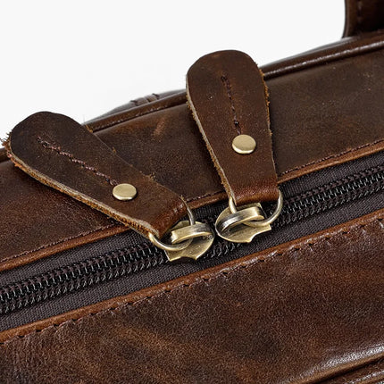 Close-up of the Leather 16" Briefcase made from genuine dark brown cow leather, showcasing the zipper and two leather zipper pulls adorned with brass buttons. The briefcase's leather exhibits visible stitching and a slightly worn texture, emphasizing its rustic and vintage appeal. An adjustable strap enhances its functionality.