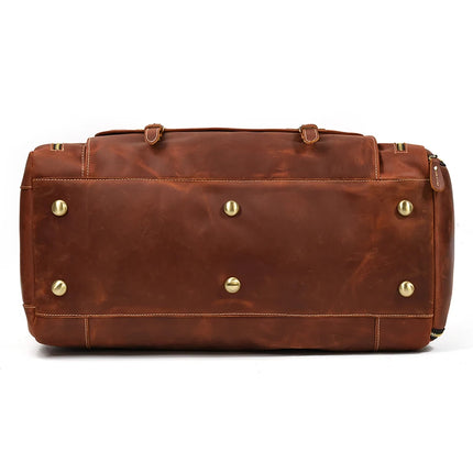 The image shows the bottom view of the Weekender 23 Leather Travel Bag made from genuine cowhide leather, featuring brass-colored metal studs for protection and support. The slightly worn leather has visible seams and a zipper on the right side, with two leather handles partially visible at the top.
