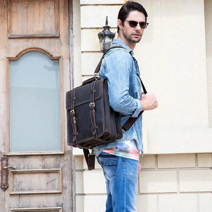 A person wearing sunglasses, a denim shirt, and jeans stands next to a vintage wooden door. They are carrying the Leather Business Travel Briefcase Backpack made of genuine cowhide leather and are looking sideways towards the camera. The background features a cream-colored wall with a lantern.