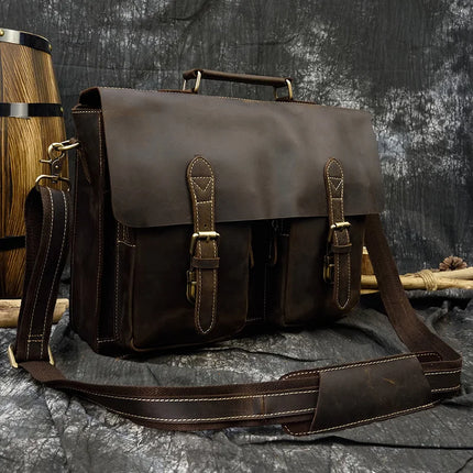 A Leather 16" Business Briefcase made of dark brown genuine cowhide leather is placed against a rugged, textured backdrop with a wooden barrel. The briefcase features a handle on top, a long adjustable strap, and two front pockets secured with metal buckles, giving it a vintage, rustic appearance.