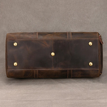 A close-up view of the bottom of a genuine cowhide **Weekender Duffle 22 Leather Travel Bag** with brass studs on a gray background. The leather has a slightly worn look, and the brass studs are placed evenly across the base for support and stability.