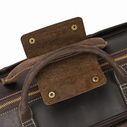 A close-up image of the genuine cowhide leather handle on the Weekender 22 Leather Travel Duffle Bag. The handle and flap cover are crafted from brown leather with visible stitching. The flap features button snaps, highlighting its secure fastening ability. A zipper segment is also visible in the background, indicating the bag's travel-friendly duffle design.
