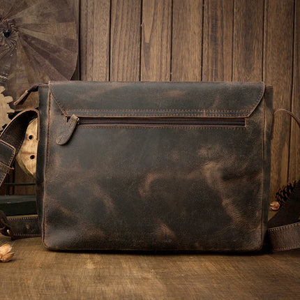 The **Vintage Leather Shoulder Bag** in brown rests on a wooden surface with a matching backdrop. Crafted from genuine cow leather, it boasts a front zippered pocket, a flap closure, and an adjustable strap with metal clasps. Its distressed, worn appearance enhances its timeless style.