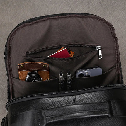 The Black Leather Travel Backpack opens to reveal a spacious interior where items are neatly organized in pockets: a wristwatch, a smartphone, a passport, and three pens. Its premium leather construction stands out against the gray surface.
