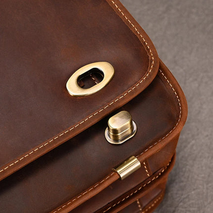 A close-up image of the Vintage Business Travel Briefcase, showcasing its brown genuine leather with visible fine stitching. The focus is on the gold-toned metal clasp and lock mechanism on the flap, which features a button-like closure and polished hardware against a plain background. An adjustable strap and spacious laptop compartment add functionality.
