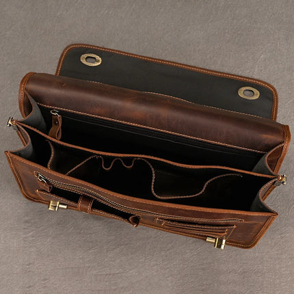 A top-down view of an open Vintage Business Travel Briefcase reveals its compartments. The brown genuine leather satchel features multiple inner pockets, including a large central compartment for a laptop, smaller zippered pockets, and additional slots for organizing items. An adjustable strap is visible against the textured gray surface.