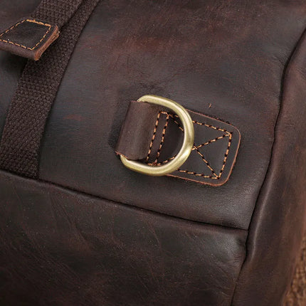 Close-up of the Vintage Leather Travel Backpack featuring a gold-tone D-ring and stitched brown strap. Crafted from genuine cowhide leather, it has a slightly distressed texture and light brown stitching. The versatile bag evokes a rugged, vintage aesthetic, perfect for the stylish traveler.