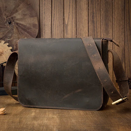 A rustic "Vintage Leather Shoulder Bag" with a timeworn look rests on a wooden surface. This genuine cowhide leather bag features an adjustable wide strap and a straightforward flap closure. The background reveals a wooden wall and an ornamental metal gear, enhancing the vintage charm.