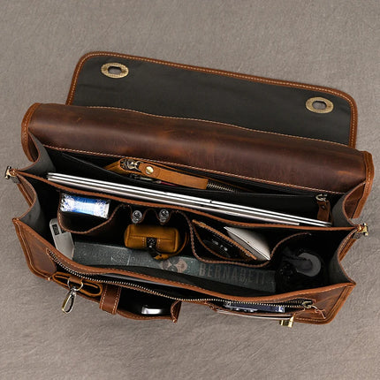 Open the brown Vintage Business Travel Briefcase made of genuine leather, featuring multiple compartments containing various items such as a laptop, book, pens, tissues, and small pouches. This satchel includes a flap with button closures and visible pockets. It also comes with an adjustable strap and a padded laptop compartment. The background surface is grey.