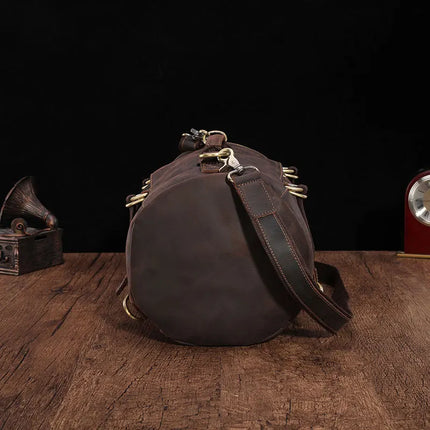 A dark brown genuine cowhide leather Vintage Leather Travel Backpack with brass hardware is displayed on a wooden surface against a black background. The stylish traveler's bag has a cylindrical shape and features a shoulder strap. Nearby, there is a vintage record player on the left and a round, red clock on the right.