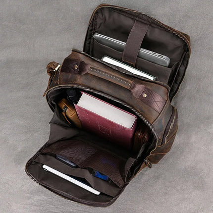 An open Vintage Leather Travel Backpack on a gray surface reveals its contents: a laptop, a tablet, a book with a red cover, a pencil case, a small bottle, and other neatly organized travel essentials in various compartments. The genuine cowhide leather enhances its timeless appeal.