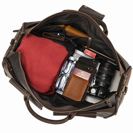 A dark Weekender 22 Leather Travel Duffle Bag is open, revealing items inside: a folded red shirt, a camera lens, a water bottle, a brown wallet, a passport, and a black notebook. Various small items are neatly organized in side compartments, and there's even a dedicated shoe compartment.
