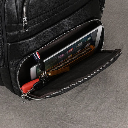 A Black Leather Travel Backpack with an open front pocket displaying various contents: a tablet, a zippered wallet, a keychain with keys, and a small red item. The zipper is halfway open, revealing the neatly arranged items inside its spacious interior. The background is a textured gray surface.