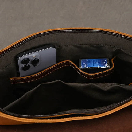 The Vintage Leather Shoulder Bag, expertly crafted from genuine cow leather and lined with black fabric, opens to unveil its interior through an unzipped top. Inside, a compartment holds a smartphone equipped with a triple camera setup on the left, while a blue packaged item is situated on the right. This bag exudes timeless style and elegance.