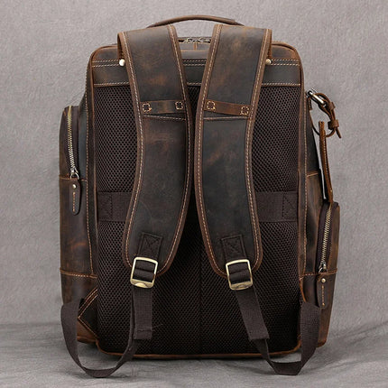 The Vintage Leather Travel Backpack, made from dark brown genuine cowhide leather, is displayed against a gray background. The back of the bag features padded straps with stitching details and adjustable buckles. Side pockets with zippers are also included, perfect for your travel essentials.