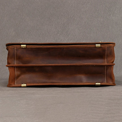 The Vintage Business Travel Briefcase is shown from the bottom view, revealing a flat base with slight creases and dark edges. The bag boasts metal corner protectors for enhanced durability. The surface showcases the texture and color variations characteristic of genuine leather, accompanied by an adjustable strap for convenience.