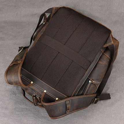 The Vintage Leather Travel Backpack, crafted from genuine cowhide leather, rests on a grey surface. It features adjustable shoulder straps and padded, breathable mesh sections on the back for added comfort. The side zippers enhance its rugged and durable design, making it perfect for carrying your travel essentials.