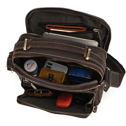 The Dark Brown Leather Shoulder Bag, crafted from genuine cowhide leather with beige stitching and brass alloy hardware, is open to reveal its contents: a wallet, smartphone, cylindrical container, keychain, tablet, blue pack of tissues, and a red case. The bag features multiple zippered compartments for organized storage.