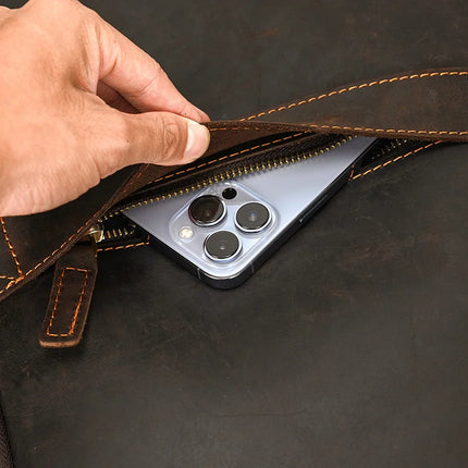 A hand is unzipping the Vintage Leather Daypack to reveal a modern smartphone with multiple camera lenses, partially visible inside the bag. This unisex backpack features genuine cowhide leather, detailed stitching, and a metallic zipper.