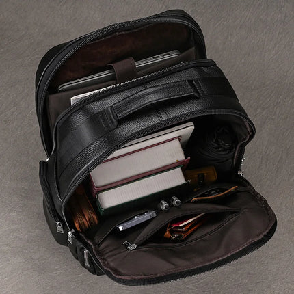 A Black Leather Travel Backpack rests open on a gray surface, revealing its roomy interior and premium leather construction. Inside the bag, you can see a laptop, two hardcover books, a notebook, an umbrella, a watch, a smartphone, earbuds, and a wallet. The backpack also features multiple compartments and zippers for optimal organization.