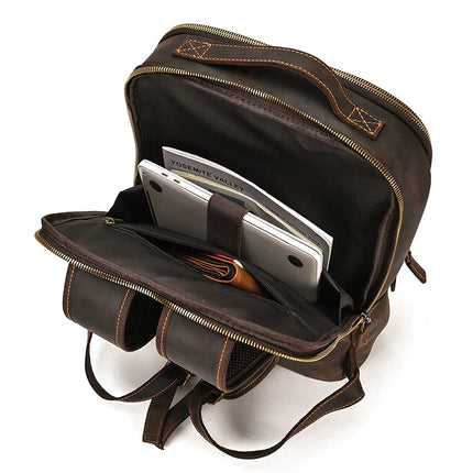 The Vintage Leather Daypack is open, revealing its contents. Inside, there are a laptop and a notebook labeled "Yosemite Valley." This genuine cowhide leather backpack has an organized interior with various compartments and is equipped with sturdy zippers and straps.