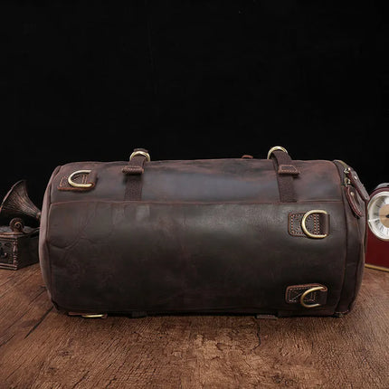 A versatile bag, the Vintage Leather Travel Backpack is crafted from genuine cowhide leather and lies flat on a wooden surface against a black background. The backpack features brass buckles, rivets, and straps for fastening. In the corner, an old-fashioned clock and gramophone are partially visible.