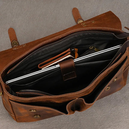 Top view of an open Leather 14" Briefcase Shoulder Bag revealing its contents. Inside, there is a laptop, a black pouch, and a small brown case. The briefcase has multiple compartments and pockets, featuring a brass snap closure. The genuine cowhide exhibits quality craftsmanship against the textured grey background.