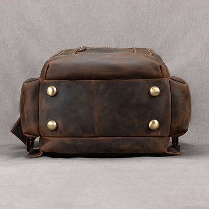 The Vintage Leather Travel Backpack is displayed from the bottom perspective against a neutral background. The base, crafted from genuine cowhide leather, features four round brass studs for support. There are also additional pockets visible on the sides to hold your travel essentials.
