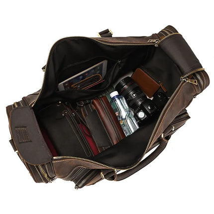 An overhead view of an open genuine leather bag filled with various items. Visible contents include a tablet in a front sleeve pocket, a camera, a water bottle, a wallet, and a book along with other small essentials. The Luggage 24 Leather Travel Bag has multiple zippers and compartments.