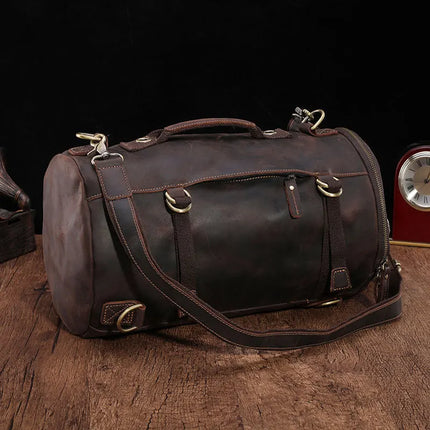 A versatile vintage-style dark brown duffle bag, known as the Vintage Leather Travel Backpack, made from genuine cowhide leather with brass hardware and a shoulder strap, is placed on a wooden surface. The background is dark, and a small red clock with a silver face is partially visible nearby. Perfect for the stylish traveler.