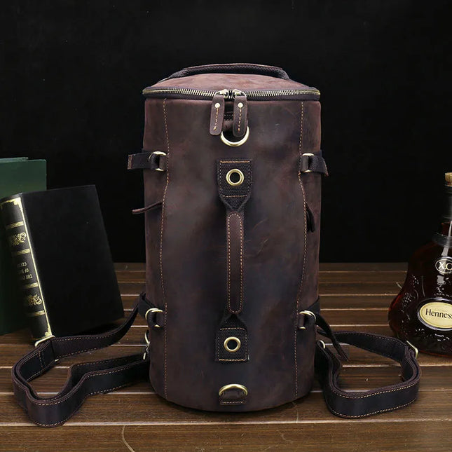 A Vintage Leather Travel Backpack, made from authentic cowhide leather and accented with brass hardware, sits on a wooden surface. This versatile backpack includes multiple straps and pockets. In the background, a couple of black and green books and a bottle of Hennessy complete the scene for the stylish traveler.