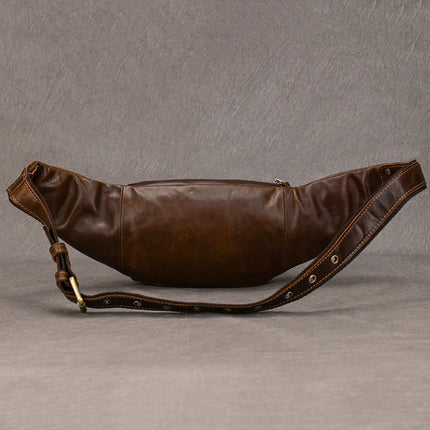 A Leather Belt Bag made from brown genuine cowhide leather is showcased against a grey backdrop. Approved by fashion experts, the bag features a polished finish, a front zipper pocket, and an adjustable strap with metal grommets and a brass buckle. The smooth leather texture displays slight creasing.