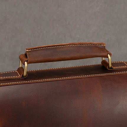 Close-up of the handle on the Vintage Business Travel Briefcase, highlighting its brass hardware connectors. The genuine leather boasts a rich, dark brown finish with visible stitching along the edges. An adjustable strap extends down to reveal part of the bag's laptop compartment against a plain grey background.