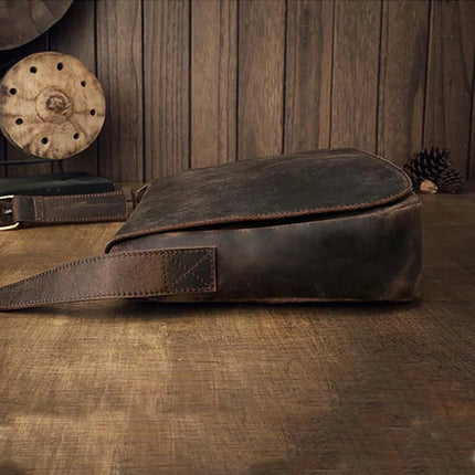 The "Vintage Leather Shoulder Bag" with a long strap rests on a rustic wooden table. Made from genuine cowhide leather, it promises durability and timeless style. The background features a wooden wall adorned with assorted decorative items, including a wooden spool and pine cones, exuding a rugged aesthetic.
