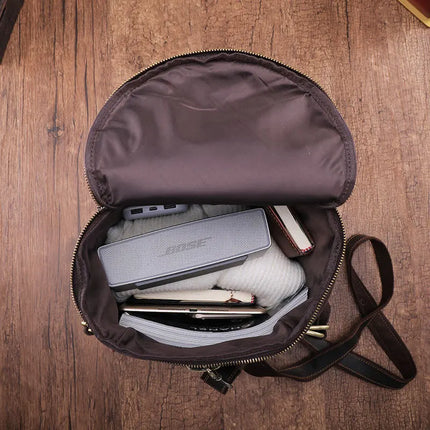 A Vintage Leather Travel Backpack crafted from genuine cowhide leather rests elegantly on a wooden surface. This open, round brown bag reveals its contents: a gray Bose speaker, a white garment, a phone, and a black device. The lid is flipped open to showcase its versatility. Complete with an attached black strap, it’s the perfect accessory for the stylish traveler.