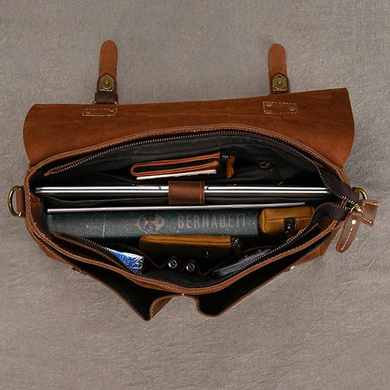 The Leather 14" Briefcase Shoulder Bag, made from genuine cowhide, is displayed open against a gray background, revealing a variety of neatly organized items inside. These include a closed laptop, a book, a pencil case, a notebook, some electronic gadgets and other miscellaneous items. The bag features two leather straps with buckles for secure closure.