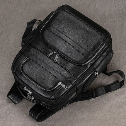 The Black Leather Travel Backpack rests on a gray surface, showcasing its premium leather construction and multiple zippered compartments. The bag features silver zippers, padded shoulder straps, and a sleek, professional design ideal for travel or daily use.