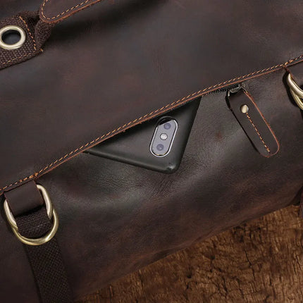 A dark brown Vintage Leather Travel Backpack made from genuine cowhide leather, featuring metal buckle accents, partially revealing a smartphone with a dual camera setup peeking out from a small pocket. This versatile bag for the stylish traveler is resting on a textured wooden surface.