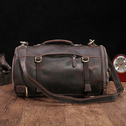 A dark brown Vintage Leather Travel Backpack, crafted from genuine cowhide leather and featuring a shoulder strap and handles, is placed on a wooden surface. The versatile backpack has a cylindrical shape, adorned with brass hardware and tan stitching. It includes pockets and buckled straps for added detail. In the background, a vintage clock is partially visible.