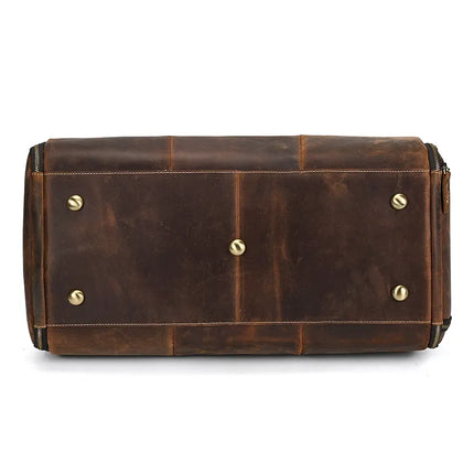 The bottom view of the Luggage 22 Leather Travel Duffle Bag showcases its antique finish on genuine leather. The base features four brass-colored metal feet for stability, divided into sections by stitched seams. Reinforced edges ensure durability, and a zipper is visible on one side, complemented by a removable strap.