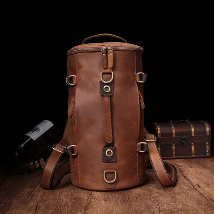The Vintage Leather Travel Backpack, a tall and cylindrical bag made from genuine cowhide leather featuring multiple buckles and straps, sits on a wooden surface. The backpack's rich brown leather exudes a vintage and worn aesthetic that makes it perfect for the stylish traveler. The background is dark, with a partially visible wine bottle and small wooden chest nearby.